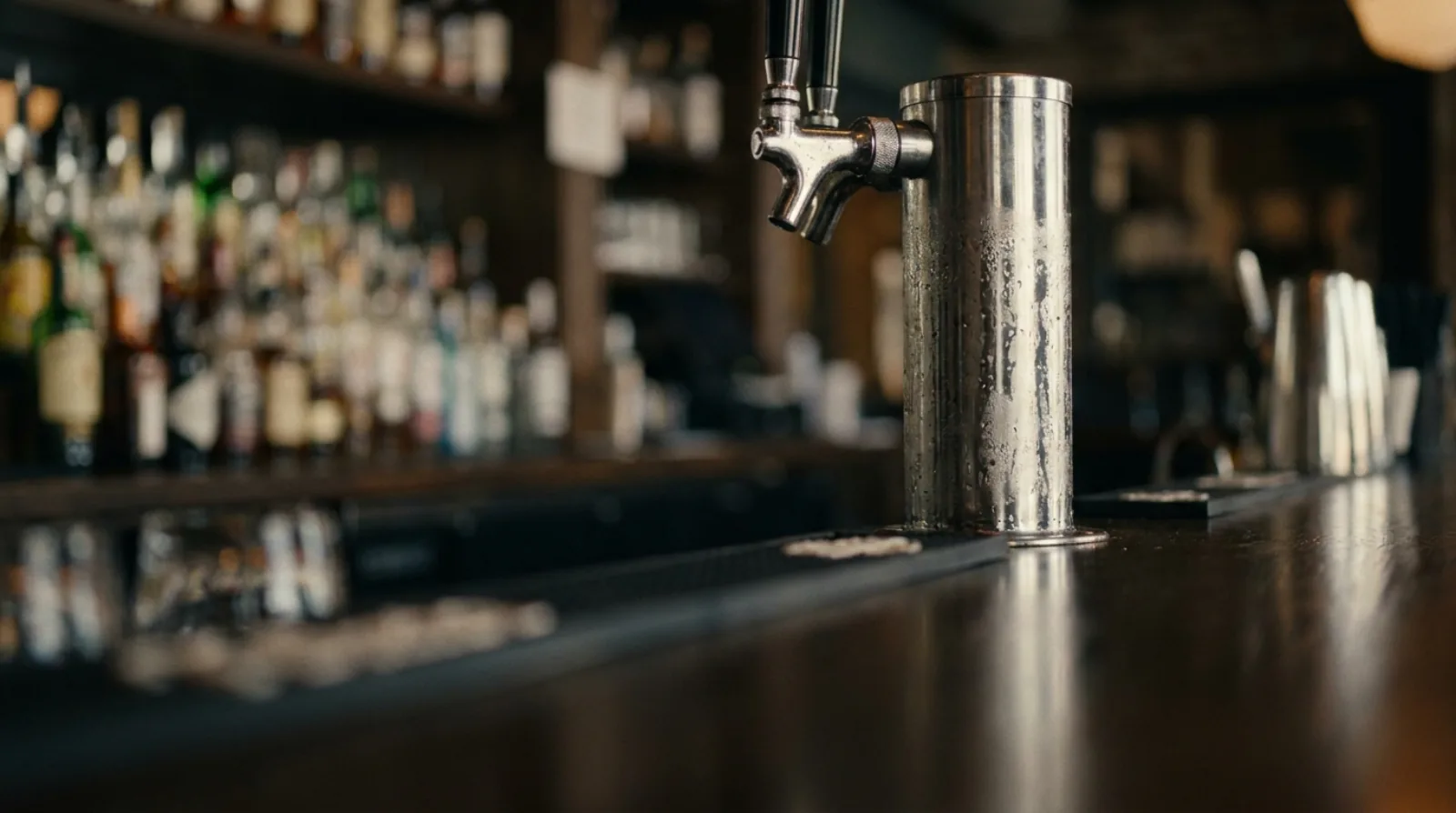 Stainless steel draft beer tap tower on a dark bar top, with a blurred bottle wall behind.